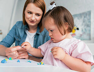 Une femme aide une petite fille trisomique dans son activité manuelle - Agrandir l'image (fenêtre modale)
